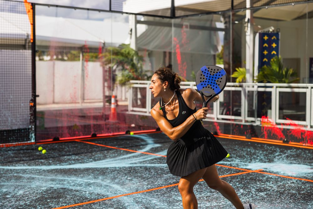 A woman in a black tennis dress lines up a backhand on an outdoor padel court with orange court lines
