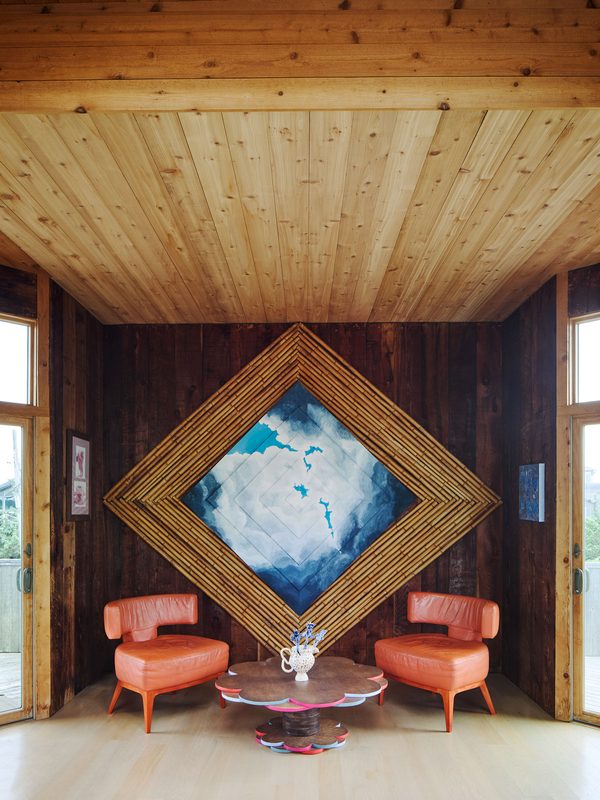 A wood-panelled room with a large painting of blue sky and clouds set in a diamond-shaped bamboo frame, above two orange armchairs and a low round table