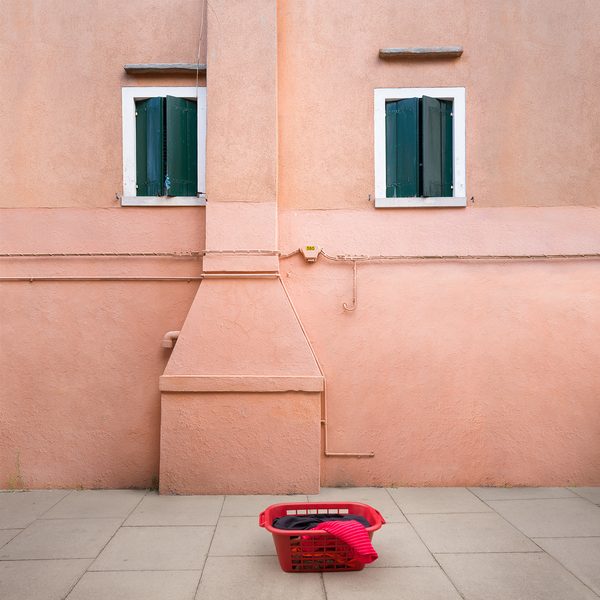 Casa Linga by Helga Stentzel: a pink house front whose two green-shuttered windows, chimney and a red laundry basket on the pavement read as a face