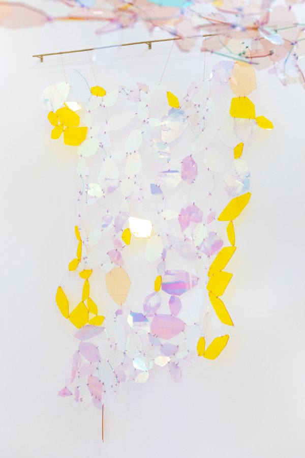 A work by Christina Watka: iridescent and yellow acrylic petal shapes stitched together with wire, hanging from a thin brass bar against a white wall